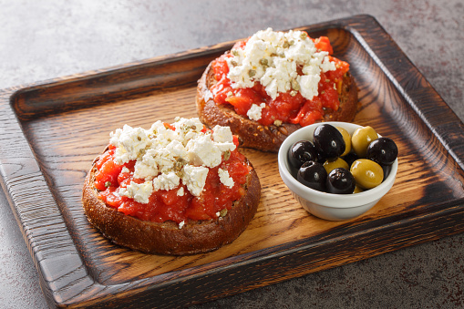 Dakos consists of a slice of cracked barley bread and garnished with crushed or grated ripe tomato and crumbled feta with oregano and olives closeup on the wooden board on the table. Horizontal
