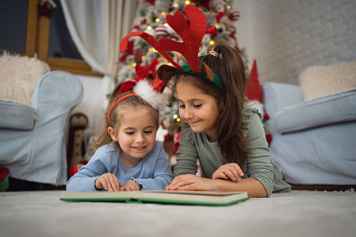 Two Caucasian sisters, lying on the floor in front of the Christmas tree and reading an fairy tale