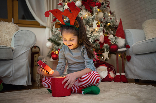 Portrait of excited Caucasian girl with costume reindeer antlers on, opening her Christmas present, while sitting in front of the Christmas