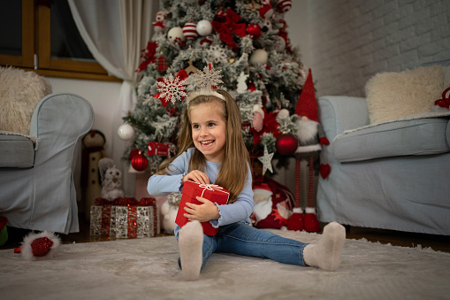 Portrait of cute Caucasian toddler girl opening her Christmas present while sitting in front of the Christmas tree