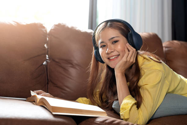 menina feliz ouvindo música e lendo o livro deitado em um sofá em casa. - room service audio - fotografias e filmes do acervo