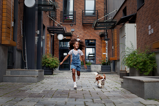 Happy black child girl with dog running on city street outside. African american little model with cocker spaniel