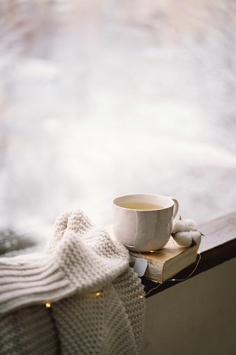 Cozy winter still life. Cup of hot tea and an open book with a warm sweater on a vintage wooden windowsill. Cozy home concept. Sweet home.
