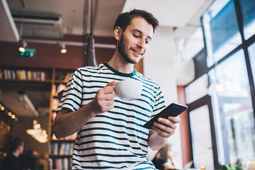 Low angle of cheerful young man with cup of hot beverage smiling and browsing smartphone while standing on blurred background of library
