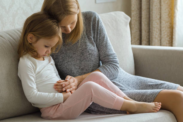 Child sitting on sofa and suffering from stomach ache at home room. Young adult mother hand touching daughter painful belly and trying calm down. Child sitting on sofa and suffering from stomach ache at home room. Young adult mother hand touching daughter painful belly and trying calm down. gastroenteritis stock pictures, royalty-free photos & images