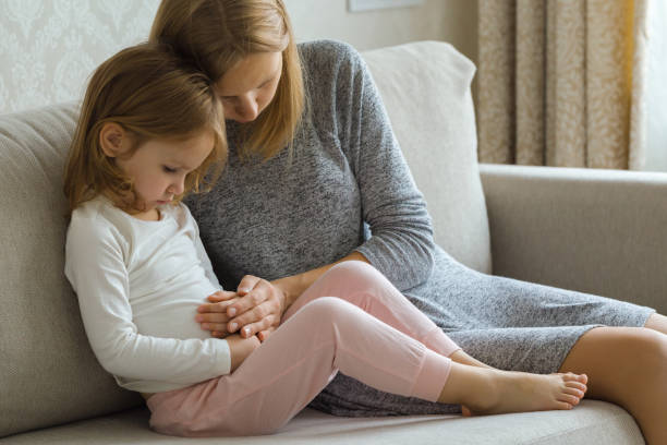 child sitting on sofa and suffering from stomach ache at home room. young adult mother hand touching daughter painful belly and trying calm down. - stomachache illness pain indigestion imagens e fotografias de stock