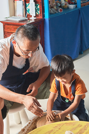 A father takes his son indoors for a pottery craft lesson, both having fun and laughing