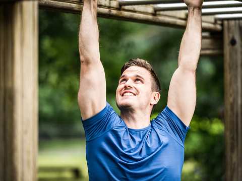 A man smiling with pride as he looks up to the last few rungs of a monkey bar at an outdoors obstacle course.