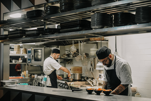 Young chef in uniform choosing fresh vegetables for dish at table with his assistant making notes on paper, they working in commercial kitchen