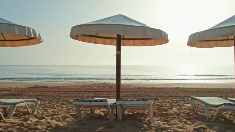 Umbrellas and beach chairs prepared for beachgoers. Sunrise. The beach in the early hours of the morning. Traces of nighttime sand raking on the beach in the morning sunshine. Lateral left camera movement. Version 4