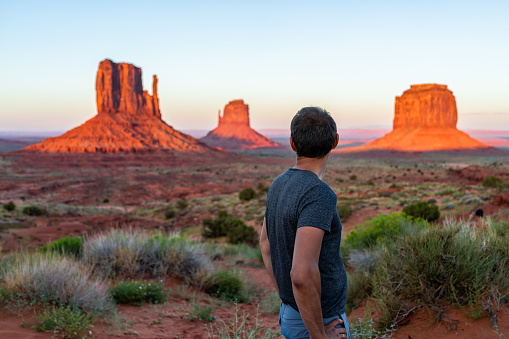 One man tourist standing looking at view of famous buttes in Monument Valley at sunset colorful light in Arizona with orange red rocks and plants