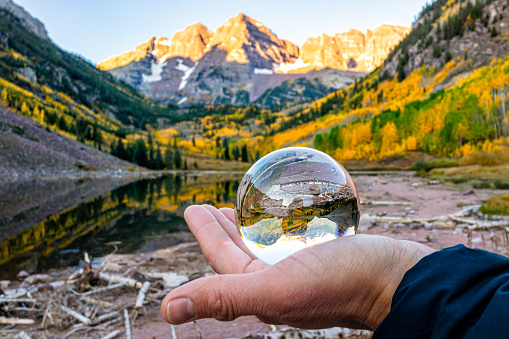Maroon Bells lake at sunrise with hand glove holding glass crystal ball reflection in Aspen, Colorado with rocky mountain peak and snow in October autumn fall season foliage