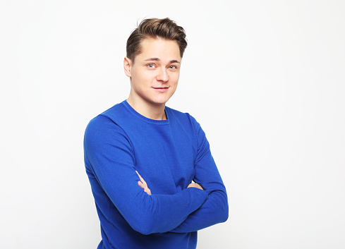 Young handsome man wearing blue sweater crossing hands and look at camera over white background