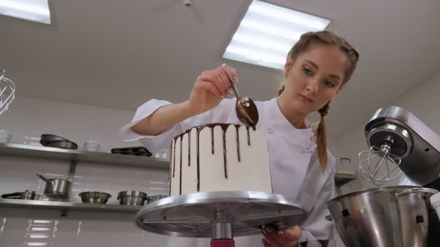 A pastry chef pouring liquid chocolate from a spoon on a white cream sponge cake