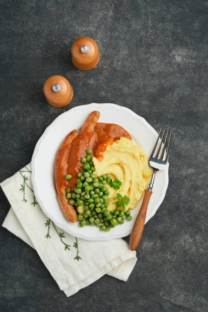 bangers und brei. gegrillte würstchen mit kartoffelpüree und grünen erbsen auf weißem teller auf schwarzem dunklem hintergrund. traditionelles gericht aus großbritannien und irland. bbq rindfleischwürste. draufsicht. - mashed potato food staple vertical color image stock-fotos und bilder