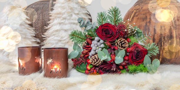 Fresh Christmas bouquet made of red roses, eucalyptus, fir branches, pine cones and other flowers lying down on a table covered with reindeer fur near two candles, feather trees and wooden decorations. Blurred lights design in front of the image.