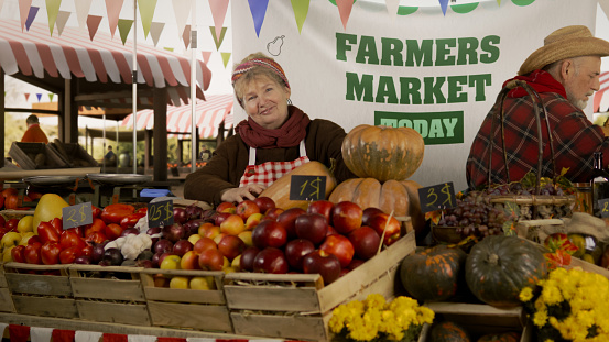 Woman farmer stands at the stall with fresh colorful fruits and vegetables, looks at camera. Farmers market festival. Vegetarian, organic and healthy food. Agriculture.