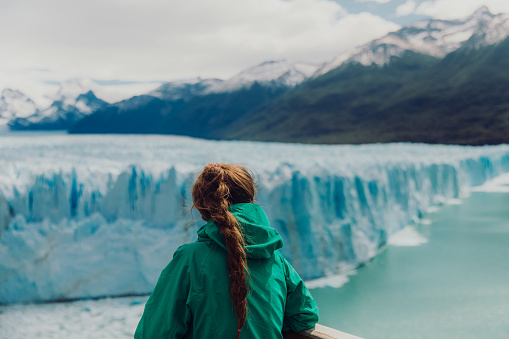Female explorer with long hair in green jacket looking at majestic lagoon with huge glacier - a part of Patagonian Ice Sheet in Argentinian Patagonia