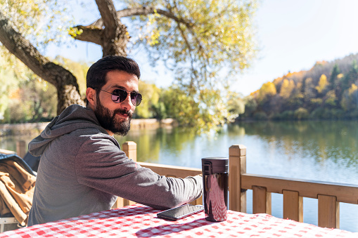 man sitting by the lake drinking coffee in autumn season. There is a lake and trees with yellowed leaves in the background. Shot with a full frame camera.