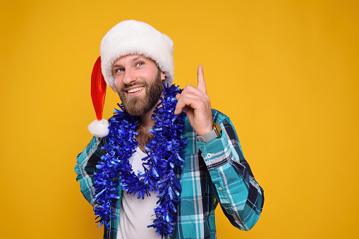 attractive young bearded man in a santa hat and with a blue boa smiles and shows that he had a great idea on a yellow background