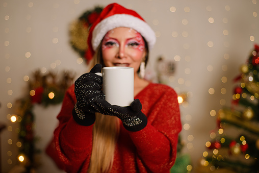 Young woman with Christmas fantasy makeup holding white mug. Selective focus on the mug. Christmas background.