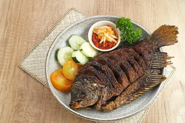 Gurame Bakar Madu, Grilled Gourami with honey and soy sauce. Served with sambal (chilli sauce), cucumber and tomatoes in white plate.