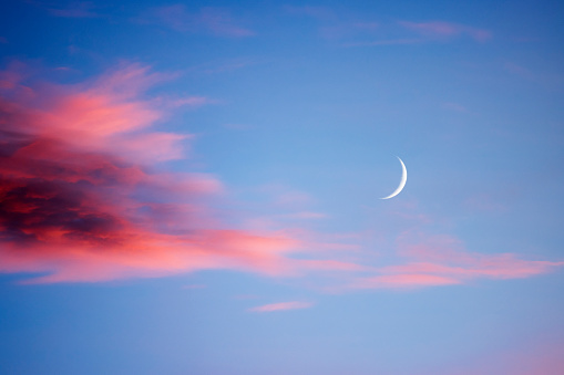 New moon at sunset with light clouds in various shades of red and magenta, darker to the left  and a blue sky behind.