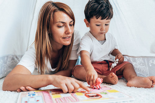 Mother and her child reading book on the floor at home. Cute little toddler with his mum reading book in child's room. Mother and son time storytelling