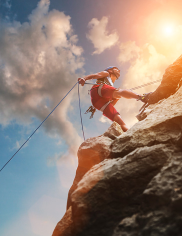 Muscular climber man in protective helmet abseiling from cliff rock wall using rope Belay device and climbing harness on evening sunset sky background. Active extreme sports time spending concept.