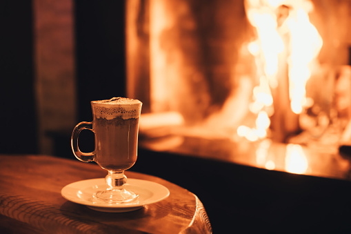 Hot chocolate in a glass on wooden table with cozy fireplace flame on the background. Winter season.