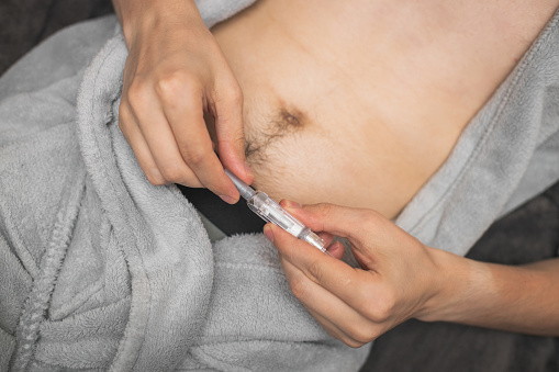 Step 3.Hands of a young caucasian man in a gray robe with a bared belly are holding an opening syringe with a medicinal liquid for an injection,lying on a bed,side view close-up.Concept step by step instructions for making an injection.