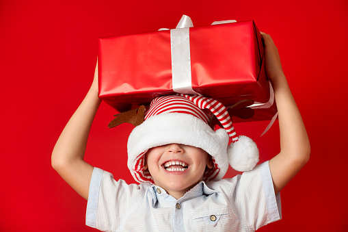 Boy laughs, holding box of gift on his head with his eyes closed on red background. Funny child in striped gnome hat sliding over his eyes and box in his hands above head