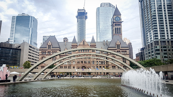 view to the Old City Hall of Toronto, modern buildings behind, with cloudy sky. the Arch and Fountain at Nathan Phillips Square in front.