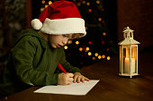 Boy in hat writes letter to Santa Claus by the light of lantern at the Xmas tree