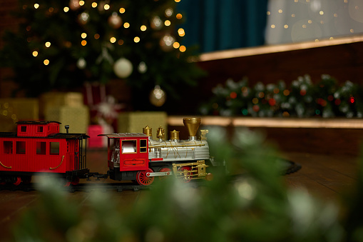 Vintage train is on the floor under a decorated Christmas tree on the background of a garland of bokeh lights. The concept of celebrating Christmas and New Year.
