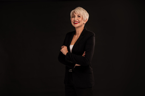 Cheerful happy businesswoman with short hairstyle and glamour makeup, looking at camera, smiling, standing against black studio background. Copy space. Success.