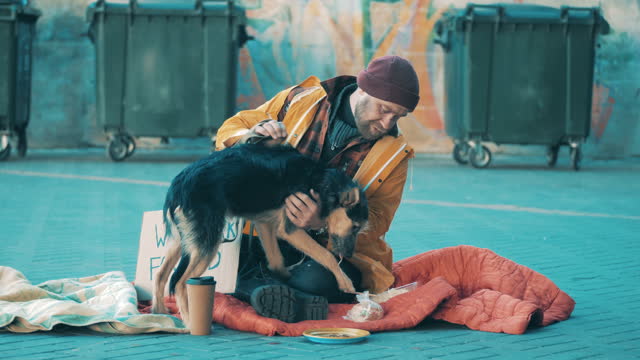 A tramp is petting his dog while sitting on the ground