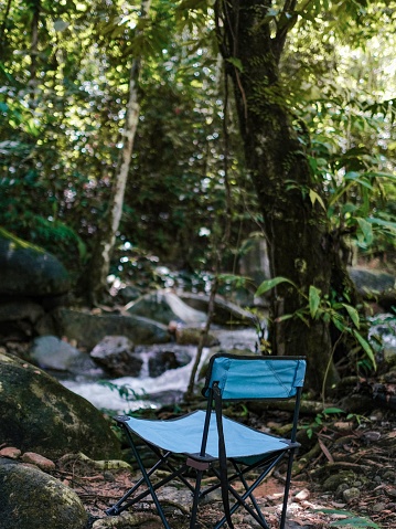 A vertical shot of an empty blue foldable camping chair in a forest, facing a water stream.