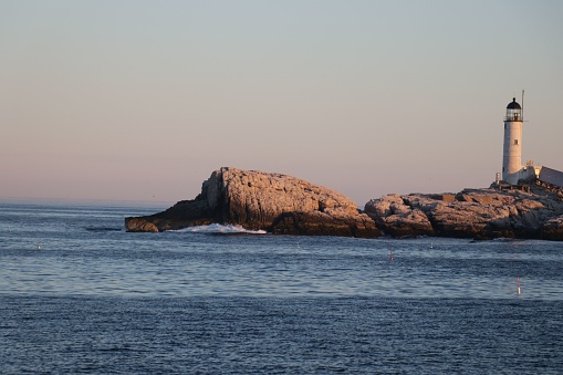 A sunset view of the Isles of Shoals Lighthouse on rocks surrounded by blue sea waves
