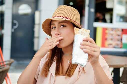 A beautiful Hispanic female in a hat eating a burrito in a cafe on a sunny day