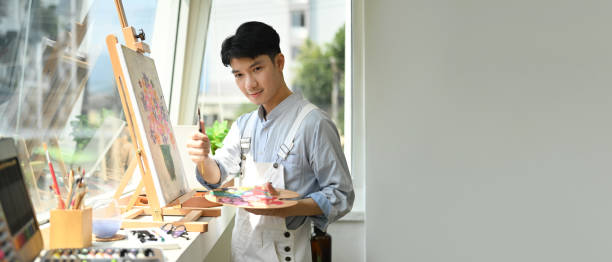 istock Asian man artist holding palette and paint brush standing in bright art studio. Panoramic web banner with copy space on the right. 1446438338