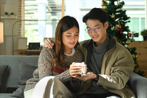 istock Romantic asian couple relaxing on comfortable couch, drinking hot chocolate and enjoying Christmas morning at home. 1446438563