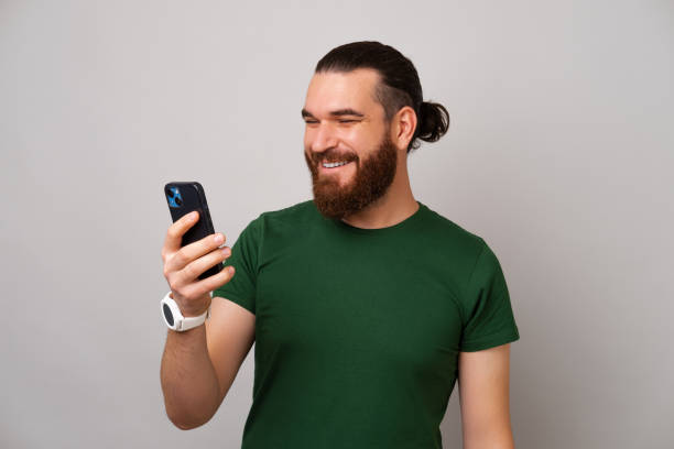 Happy bearded man is holding his phone and looking at it while misling.