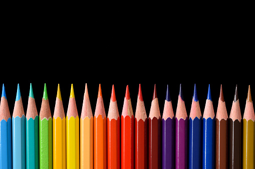 Color image depicting a close up macro view of a collection of colored pencils in a row, on a black background. Selective focus image with one of the pencils in sharp focus with the others blurred out of focus.