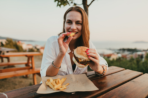 Happy woman sitting outdoors and eating burger and fries on a sunny summer day during a seaside vacation