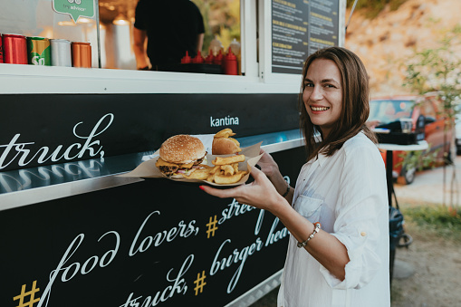 Happy young woman buying burgers and fries at the food truck during her summer vacation