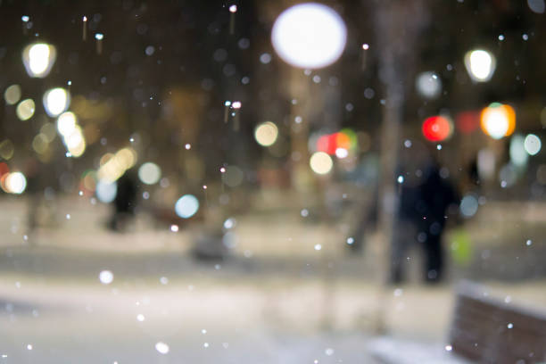 Blurred background. City view, lights, falling snow, night, street, bokeh spots of headlights of moving cars. Diffuse Urban backdrop winter scenery of street in city at night. Lantern light, snowfall