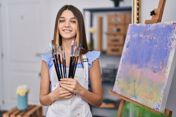 adorable fille souriante confiante tenant des pinceaux dans un studio d’art - artist child artists canvas indoors photos et images de collection