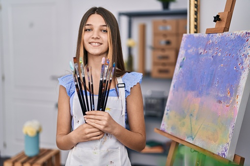 Adorable girl smiling confident holding paintbrushes at art studio