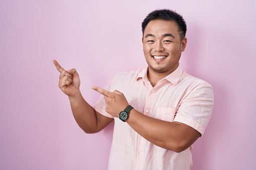 Chinese young man standing over pink background smiling and looking at the camera pointing with two hands and fingers to the side.